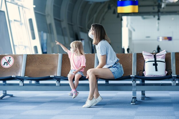 Mother with daughter in masks waiting for their flight at airport woman with little girl in