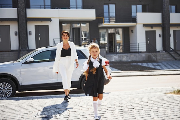 Mother with daughter in school uniform outdoors near white car.