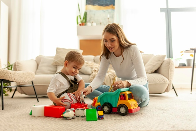 mother with her little son sits on the carpet at home and plays with toys in the room