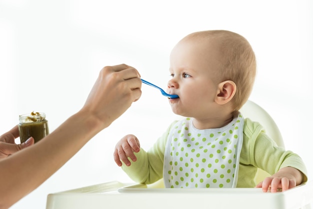 Photo mother with jar of baby nutrition and spoon feeding infant on feeding chair on white background