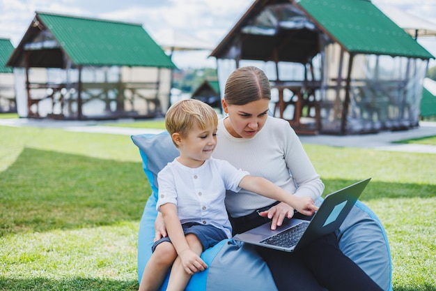 A mother with a laptop her son sitting next to her outdoors on
a green grass lawn rest and work with a child