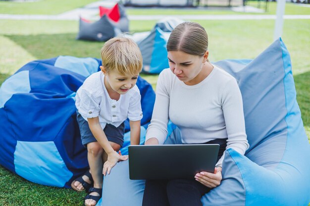 A mother with a laptop her son sitting next to her outdoors on\
a green grass lawn rest and work with a child