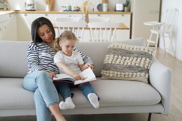 Мать с маленькой дошкольной дочерью вместе читают книгу, учатся, наслаждаются семейным хобби. Мама обнимается, воспитывает девочку, сидящую на диване, читает сказку дома. Воспитание, материнство.