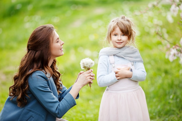 Mother with small daughter blowing to dandelion - lifestyle outdoors scene in park. Happy family concept