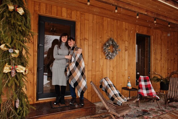 Mother with son in covered blanket in winter day against wooden house