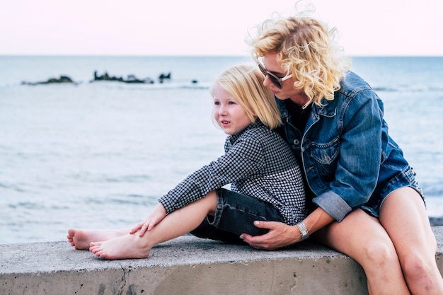 Mother with son sitting on a concrete embankment against sea. Affectionate mother caring about her child outdoors. Young woman in sunglasses spending leisure time with little boy during vacation.