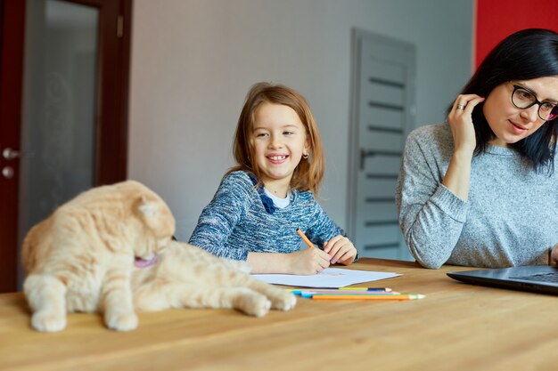 Mother working in her home office on a laptop, her daughter sits next to her and draw, Scottish cat sitting on the table too. Woman freelance, remote work and raising a child at workplace.