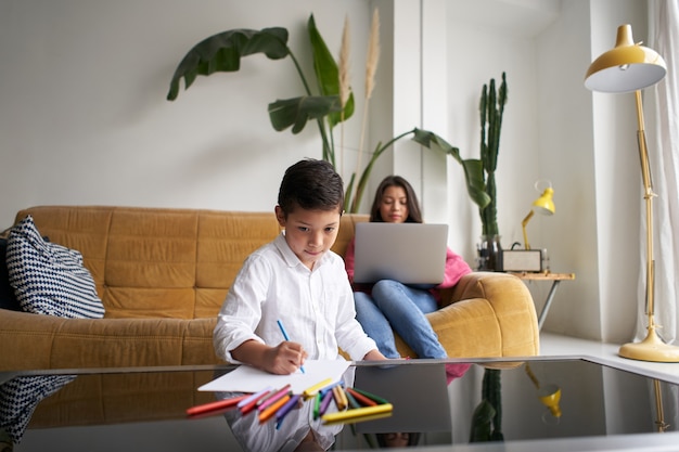 The mother works on the laptop while her son paints pictures on the living room table lifestyle with...