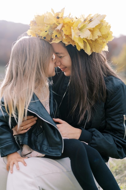 Photo mother in a wreath of yellow leaves touches noses with a little girl sitting on her lap