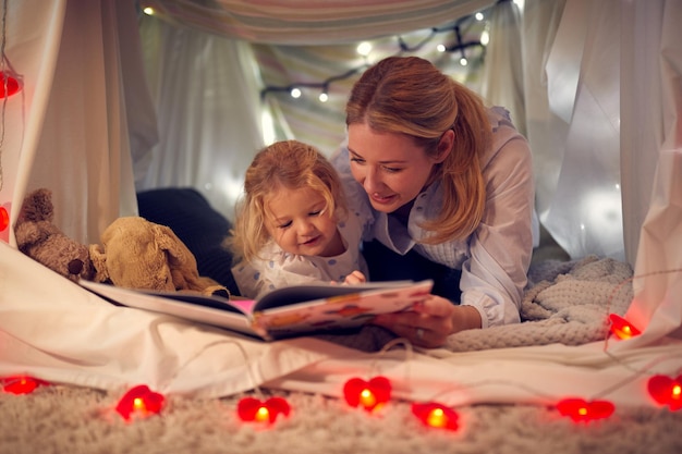 Mother And Young Daughter Reading Story In Homemade Camp In Child39s Bedroom At Home