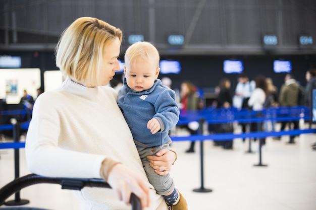 Motherat reist met zijn babyjongenskind dat duwt met een kinderwagen en een bagagewagentje op het eindstation van de luchthaven Reizen met kindconcept