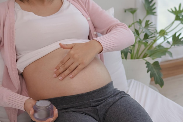 Motherhood asian young pregnant woman applying pregnancy lotion prevent stretch marks on belly by rubbing cream with moisturizer, moisture for abdomen, belly beauty skin, health care at home.