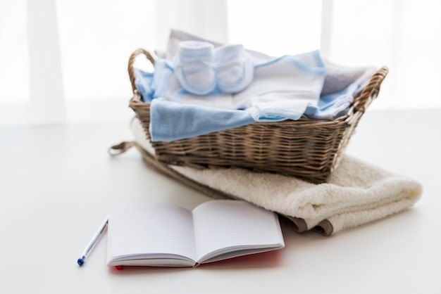 motherhood, care, parenthood and object concept - close up of pile of baby clothes with towel for newborn boy in basket and blank notebook or diary on table