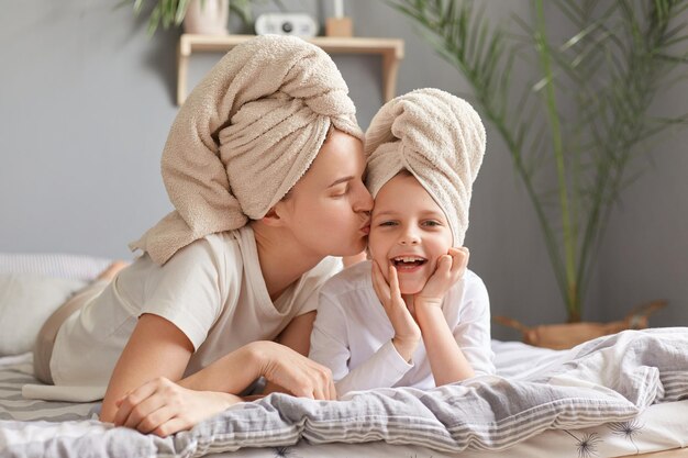 Motherhood childhood parenthood Loving mother and daughter laying in bed wearing towel posing in bedroom at home woman kissing her charming kid doing beauty procedures together