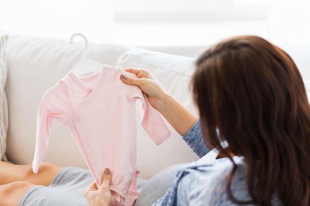 motherhood, pregnancy, people and kids clothing concept - close up of happy woman holding and looking at pink baby girls bodysuit at home