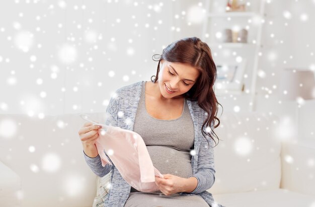 motherhood, pregnancy, people and kids clothing concept - happy woman holding and looking at pink baby girls bodysuit at home over snow