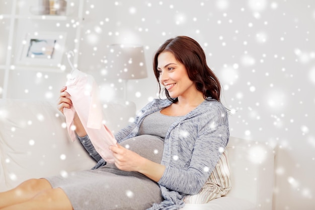 motherhood, pregnancy, people, winter and kids clothing concept - happy woman holding and looking at pink baby girls bodysuit at home over snow