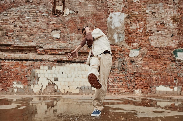 Motion shot of young male dancer posing against brick wall outdoors in shabby urban setting