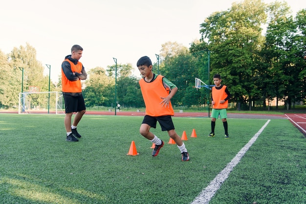 Мотивированные мальчики в оранжевых жилетах бегают среди пластиковых конусов во время тренировки по футболу на стадионе.