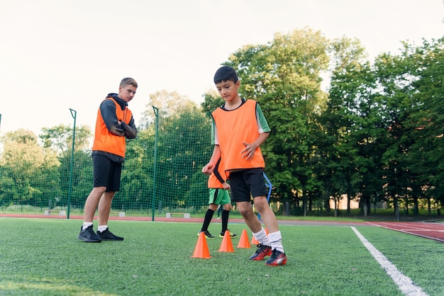 スタジアムでのサッカーのトレーニング中にプラスチックのコーンの間を走っているオレンジ色のベストを着たやる気のある男の子。