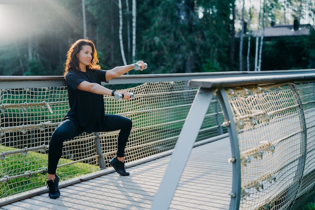Motivated brunette active young woman does squat exercises with dumbbells trains biceps dressed in black active wear poses at bridge outdoor during sunrise has morning workout Sport concept
