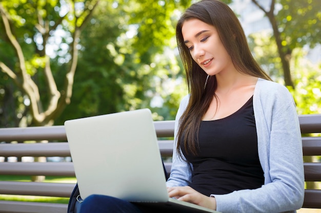 Motivated freelancer in the park. Young brunette girl on a bench, smiling and working with laptop online. Lifestyle, technology and remote job concept