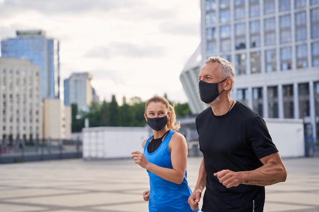 Motivated middle aged man in protective face mask running together with his wife in urban