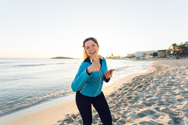 Motivated sporty woman doing thumbs up success gesture after urban workout on seashore.