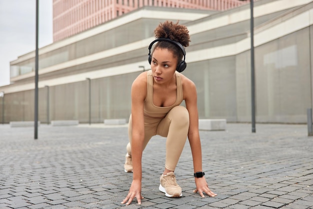 Motiva la donna sportiva pronta per correre in piedi nella posa della linea di partenza indossa cuffie wireless sulle orecchie tuta da ginnastica e scarpe da ginnastica