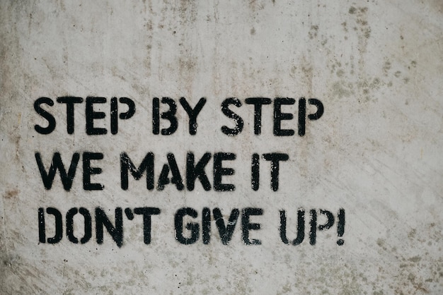 Photo the motivational quote 'step by step we make it, dont give up' written on a white wall with black paint