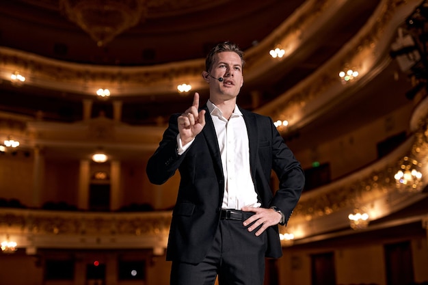 Motivational speaker in formal elegant suit, talking about\
happiness, self, success and how better more productive self. tech\
startup presenter pitching. handsome caucasian male on stage