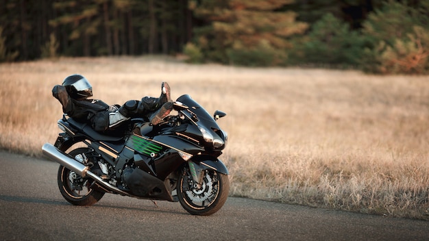 Motorcycle driver in a leather suit lies on a motorcycle alone on an asphalt road against a forest background