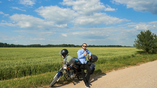 Photo a motorcycle on an empty dirt road next to a wheat field a motorcyclist wearing sunglasses eats ice cream on a hot sunny day a guy in a blue shirt and black jeans is sitting on a motorcycle