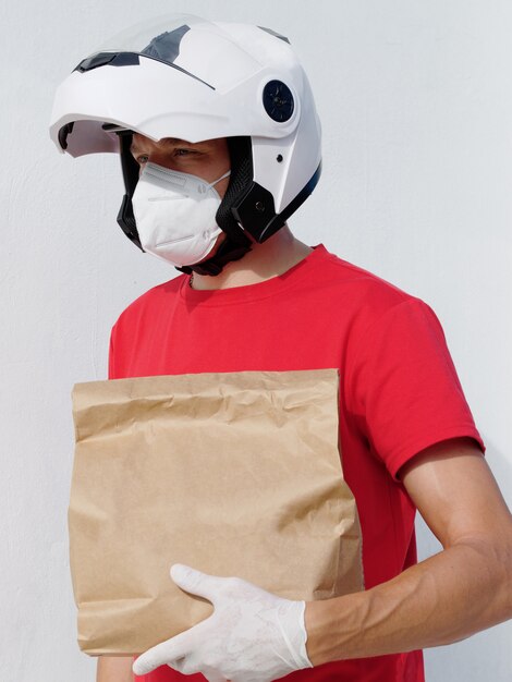 Фото Мотоциклист доставщик в униформе. держит картонную сумку в медицинских защитных перчатках и маску kn95. онлайн покупки и экспресс доставка.