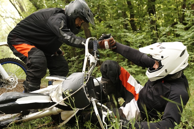 Motorcyclist in helmet helping friend fallen out of motorcycle in forest, friendship and support concept