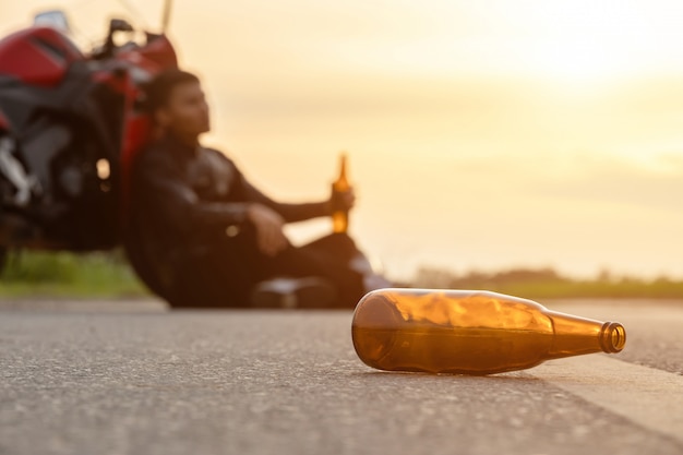 Motociclista seduto sulla strada accanto alla sua moto, bevendo un alcol o birra