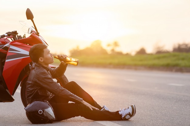 Motorcyclist sitting on the road beside his motorcycle, drinking an alcohol or beer