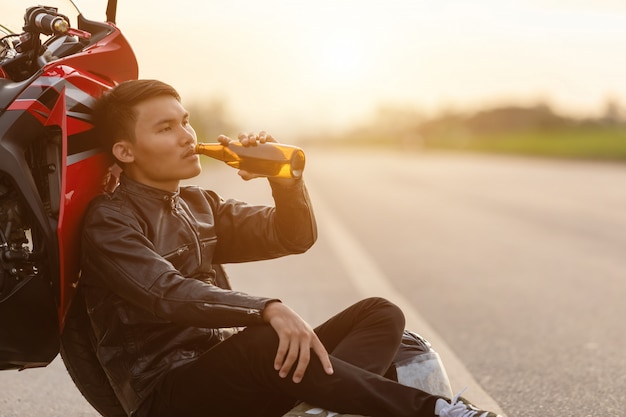 彼のバイクの横の道路に座ってビールを飲むモーターサイクリスト