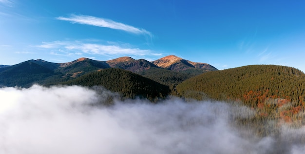 Mountain range against a blue sky. Natural landscape of mountains peaks, forest and low clouds from aerial view.
