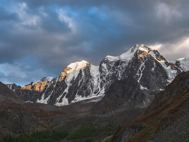 日の出の山脈。日の出の曇り空に太陽に照らされた雪の山々の素晴らしい風景。日の出の色の大きな氷河と暗い風光明媚な風景。