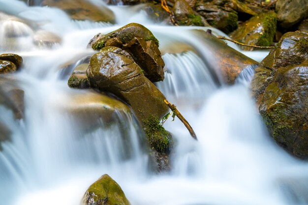 Mountain river with small waterfall with clear turquoise water falling down between wet boulders with thick white foam.