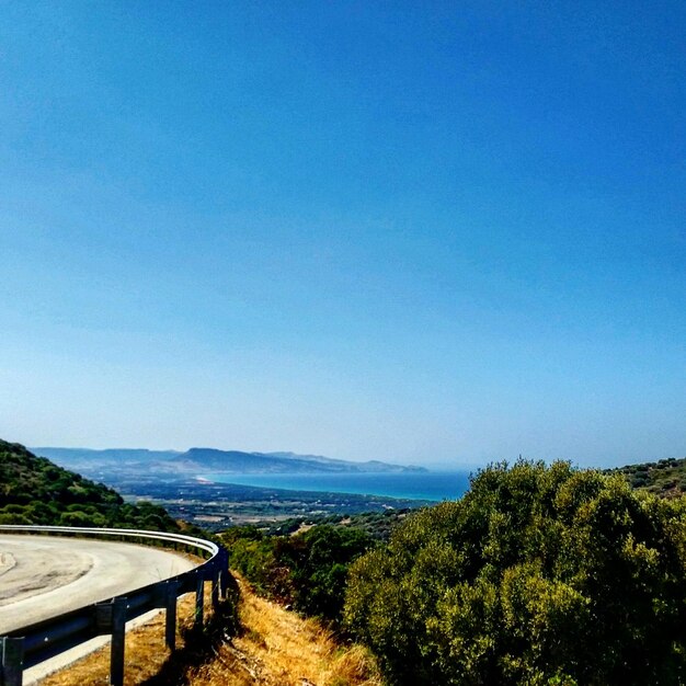 Mountain road by trees growing against clear blue sky