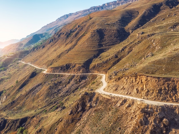 Mountain serpentine in the dawn sunlight. Dangerous narrow dirt mountain road through the hills to a high-altitude village. Aerial view.