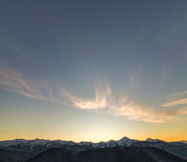 Mountain winter panorama landscape at dawn. Steep mountain peaks covered with snow and woody hills with spruce forest, raising bright sun on clear blue sky at sunrise .