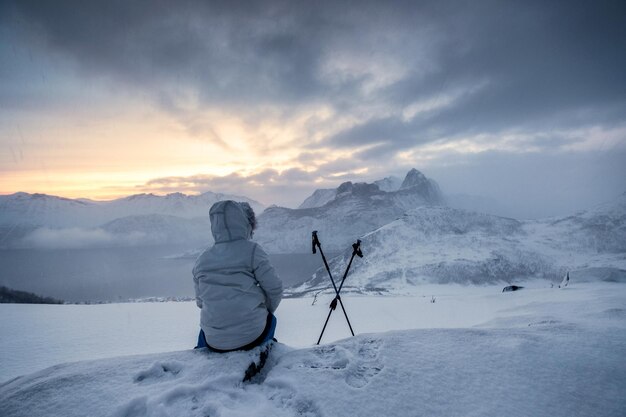 雪のピークの凍るような天気でトレッキングポールと一緒に座って日の出を待っている登山家