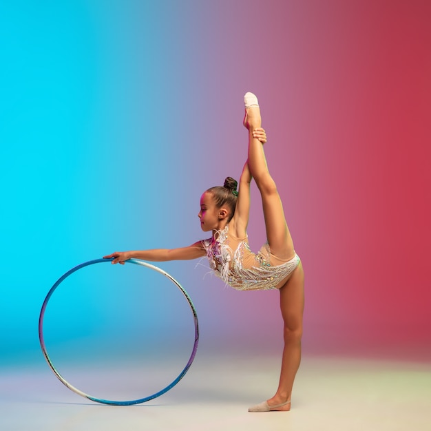 Movement. Little caucasian girl, rhytmic gymnast training, performing isolated on gradient blue-red studio background in neon. Graceful and flexible, strong child. Concept of sport, motion, action.