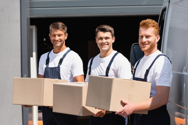 Movers in uniform holding carton boxes and looking at camera near truck on urban street