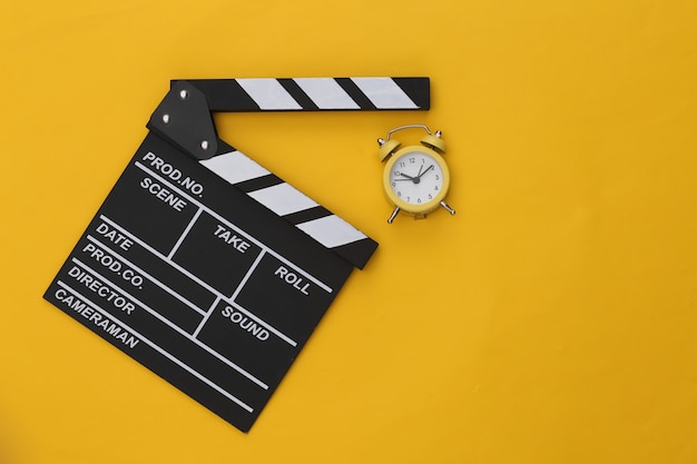 Movie clapperboard and mini alarm clock on yellow background.