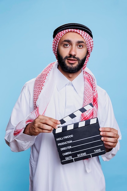 Movie scene director wearing traditional muslim clothes holding clapperboard and looking at camera Arab dressed in islamic headscarf and thobe using clapboard studio portrait