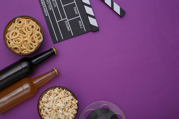 Movie time. Movie clapperboard, bottles of beer, snacks, film reel on purple background copy space. Top view